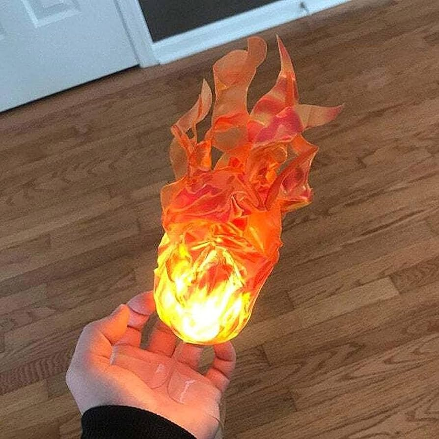 A hand holding a small flame-shaped decorative object with glowing orange and yellow faux flames, photographed over a wood floor.