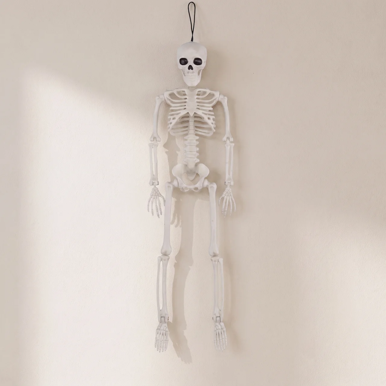 A hanging white plastic skeleton decoration with a black hanging loop at the top, shown against a plain off-white wall.