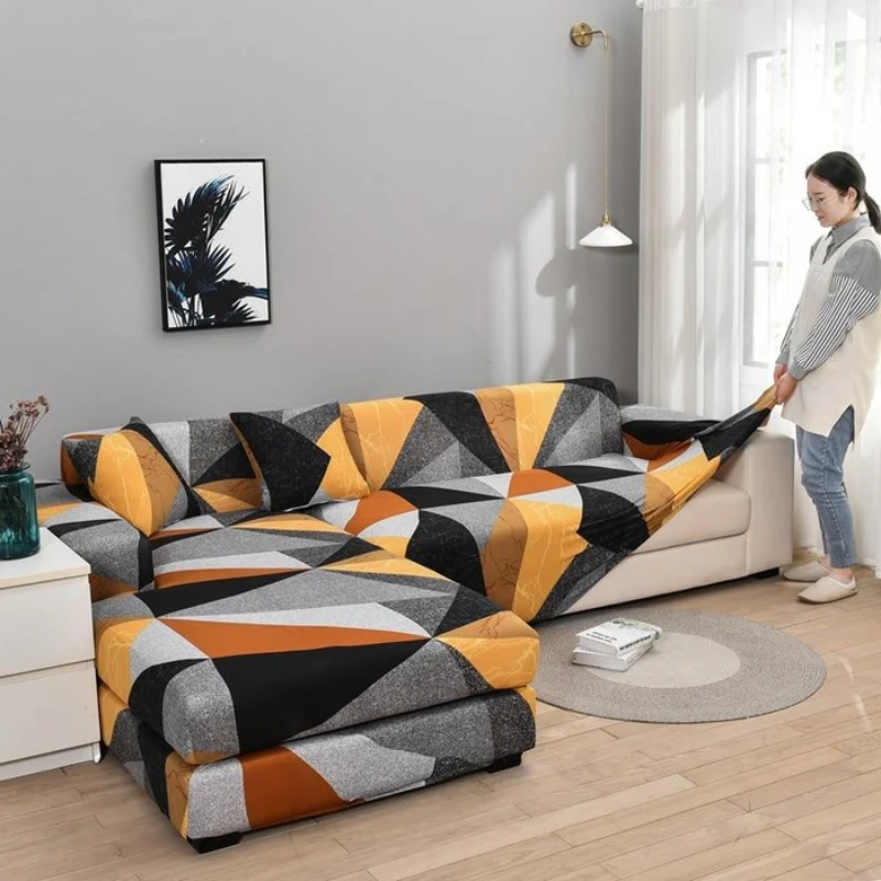 A woman in a home setting adjusts a geometric-patterned sofa slipcover on a sectional sofa. The cover features a modern abstract design in orange, yellow, black, gray, and white.