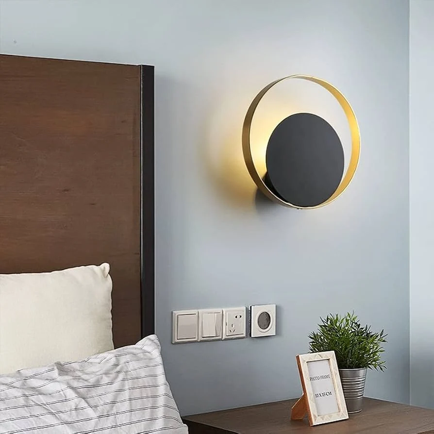 A modern circular wall sconce with a gold outer ring and a black inner disc, mounted on a light blue-gray wall above a bedside table next to a bed.