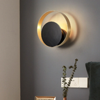 A modern wall-mounted light fixture with a gold-toned circular frame and a dark central panel, shown lit on a gray wall above a table with a vase of eucalyptus.