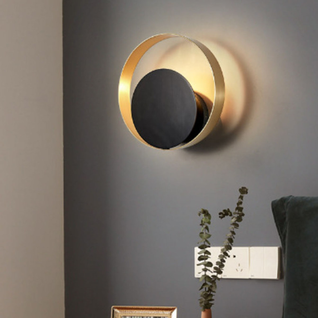 A modern wall-mounted light fixture with a gold-toned circular frame and a dark central panel, shown lit on a gray wall above a table with a vase of eucalyptus.