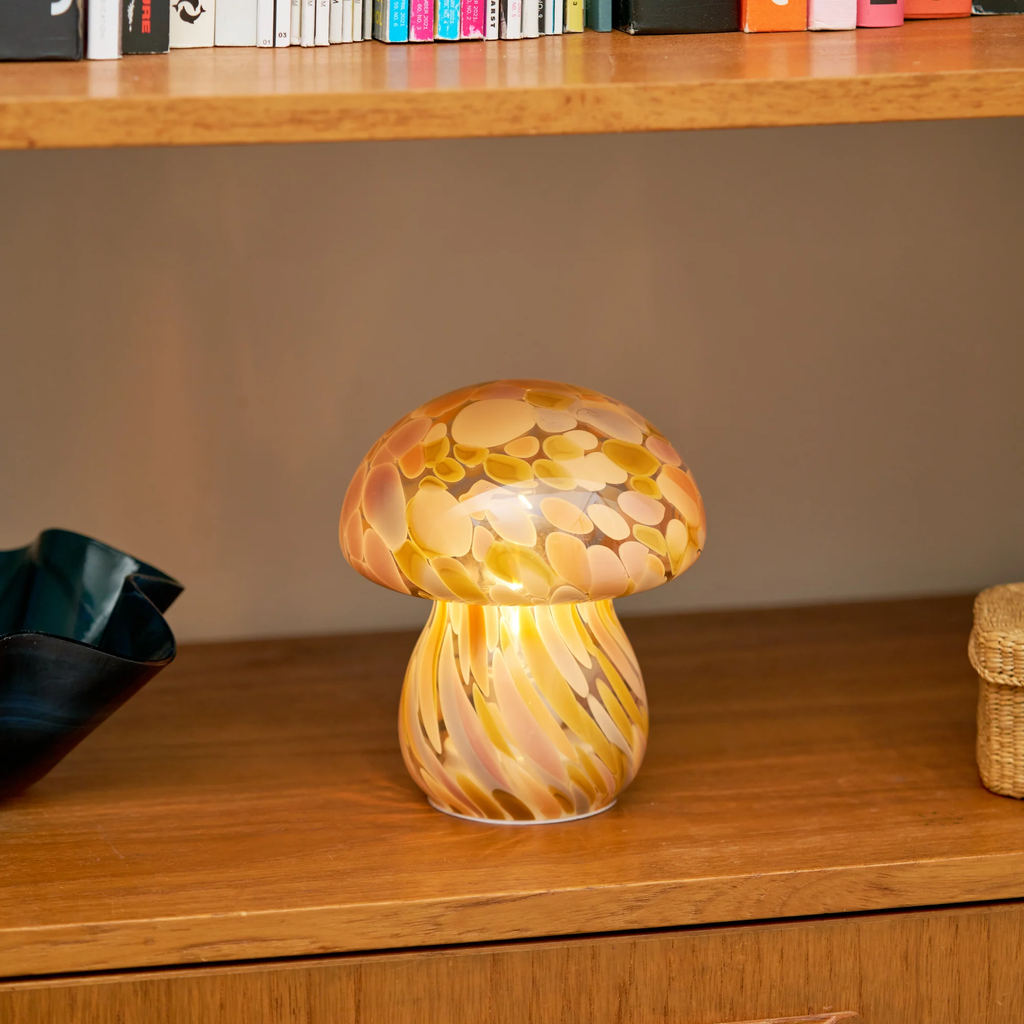 A mushroom-shaped glass lamp with a marbled pattern of yellow, orange, and cream, sitting on a wooden shelf.