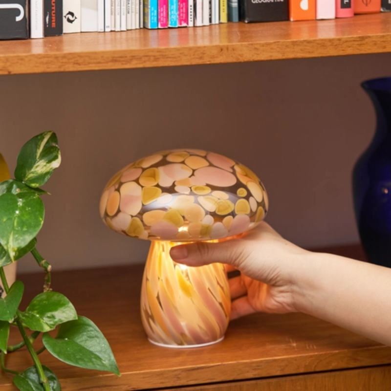 A mushroom-shaped decorative lamp with a glossy, mottled cap in shades of pink, yellow, and gold, and a striped stem glowing with warm light. A hand is touching the lamp, which is placed on a wooden shelf next to a green plant.