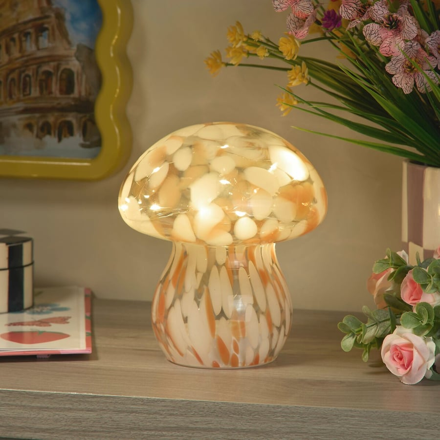 A mushroom-shaped decorative lamp or figurine with a glossy translucent cap and a marbled stem in cream, orange, and white, placed on a tabletop in a styled indoor setting with flowers and framed wall art in the background.