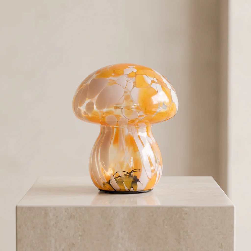 A small mushroom-shaped decorative object with a glossy orange and cream marbled cap and stem, displayed on a light stone pedestal against a soft beige background.