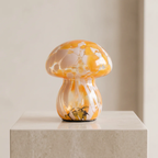 A small mushroom-shaped decorative object with a glossy orange and cream marbled cap and stem, displayed on a light stone pedestal against a soft beige background.