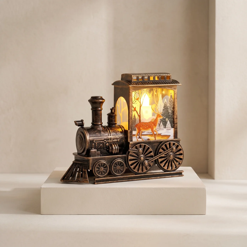 A decorative tabletop lamp shaped like a vintage steam locomotive train, displayed on a white pedestal against a warm beige background. The lamp features a dark bronze-colored metal body with a glowing warm yellow light inside a windowed cabin showing a deer and winter scene.