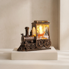 A decorative tabletop lamp shaped like a vintage steam locomotive train, displayed on a white pedestal against a warm beige background. The lamp features a dark bronze-colored metal body with a glowing warm yellow light inside a windowed cabin showing a deer and winter scene.