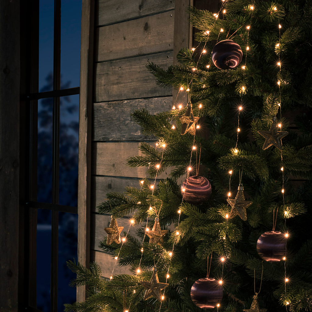 Warm white string lights draped over a decorated Christmas tree, with gold star ornaments and purple marbled baubles, set against a rustic wooden wall and window at dusk.