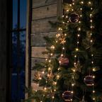 Warm white string lights draped over a decorated Christmas tree, with gold star ornaments and purple marbled baubles, set against a rustic wooden wall and window at dusk.