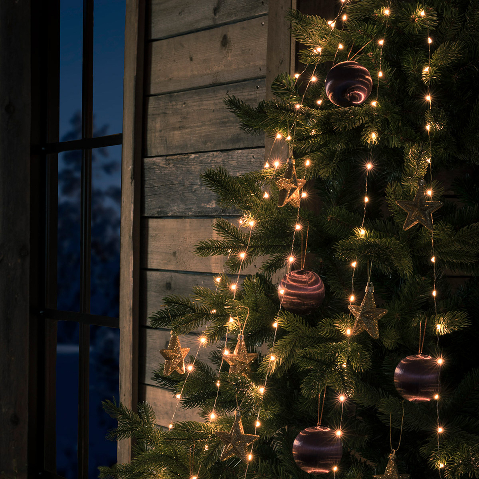 Warm white string lights draped over a decorated Christmas tree, with gold star ornaments and purple marbled baubles, set against a rustic wooden wall and window at dusk.