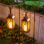 Two outdoor solar garden lanterns with warm glowing LED lights, mounted on black metal hangers in a garden setting in front of a wooden fence.