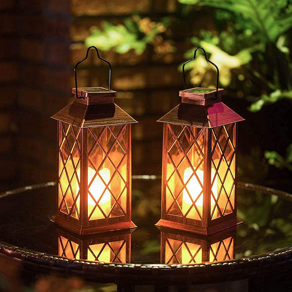 Two decorative lanterns with warm glowing candles inside, placed on a glass table outdoors at night with a blurred brick wall and greenery in the background.