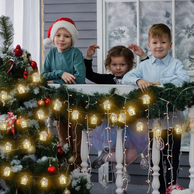 Three children posing behind a decorated white railing at a holiday house window, with glowing house-shaped Christmas string lights hanging down and a decorated Christmas tree in the foreground.