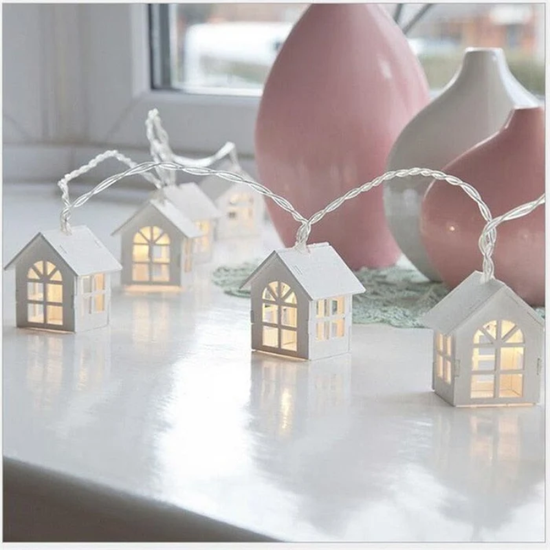 String lights shaped like small houses on a reflective surface with pink vases in the background.