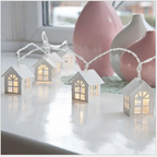 String lights shaped like small houses on a reflective surface with pink vases in the background.