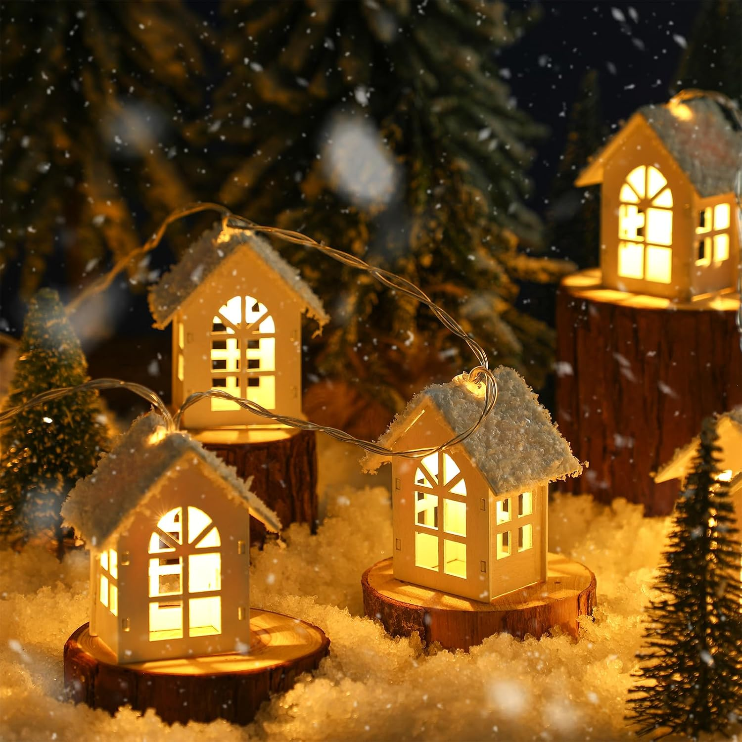 Warm white fairy lights shaped like small house-shaped lanterns, decorated with faux snow and displayed on wooden tree stumps in a snowy, festive outdoor scene.