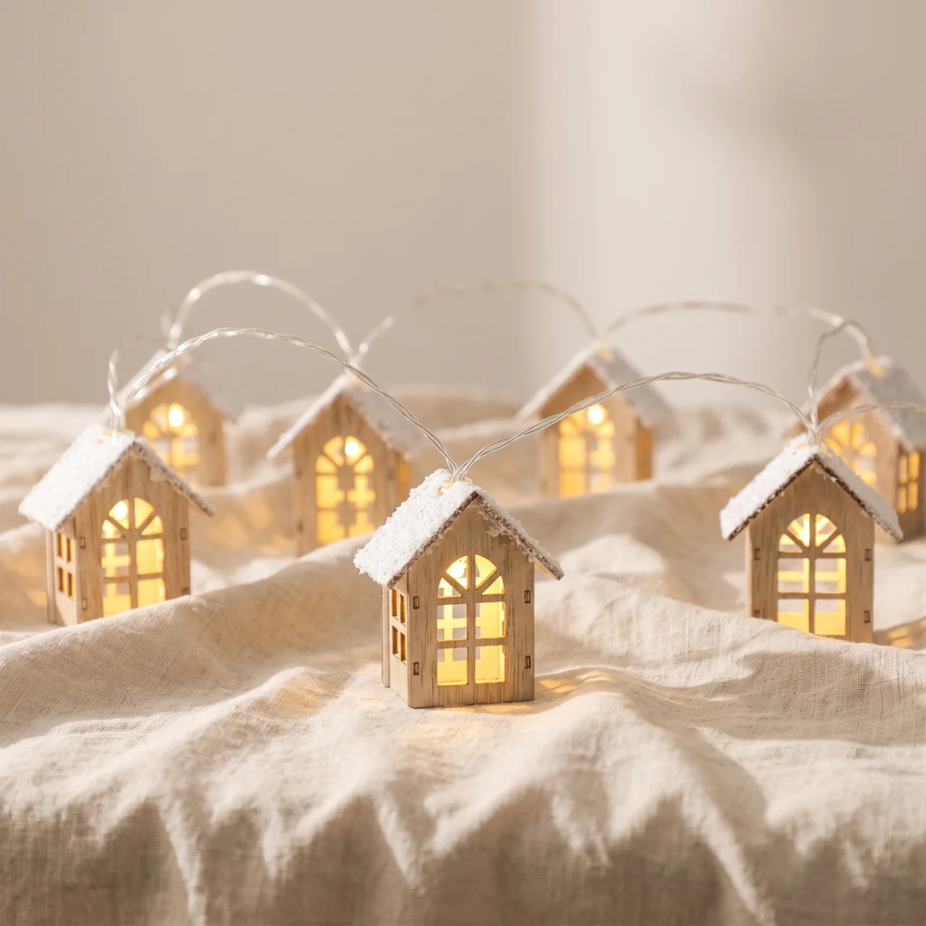 A set of miniature wooden house string lights with glowing warm yellow windows and white faux-snow on the roofs, arranged on a beige linen fabric background.
