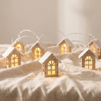 A set of miniature wooden house string lights with glowing warm yellow windows and white faux-snow on the roofs, arranged on a beige linen fabric background.