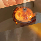 A small round diffuser or humidifier being held in two hands, glowing with warm orange light and displaying dried flower petals inside a clear dome. The scene is softly lit and styled, with a beige background and decorative floral overlay in the top left.