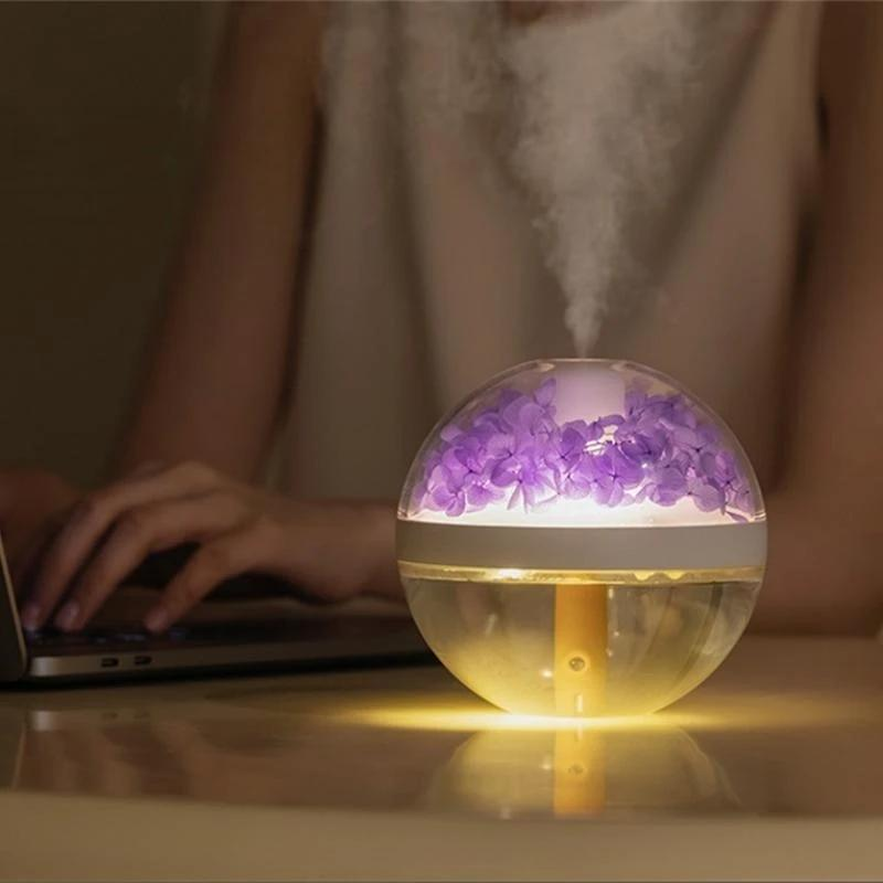 A spherical humidifier or diffuser on a tabletop, with purple flowers inside and a warm yellow glow from the illuminated interior. Steam is rising from the top, and a person's hands and torso are blurred in the background near a laptop.