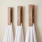 Three wooden clothespins mounted on a beige wall, each holding a white towel. The clothespins have a natural wood finish with a small silver metal spring.
