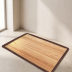 A rectangular mat with a light wood grain pattern and a dark brown stitched border, shown on a light beige floor in a minimalist room corner.