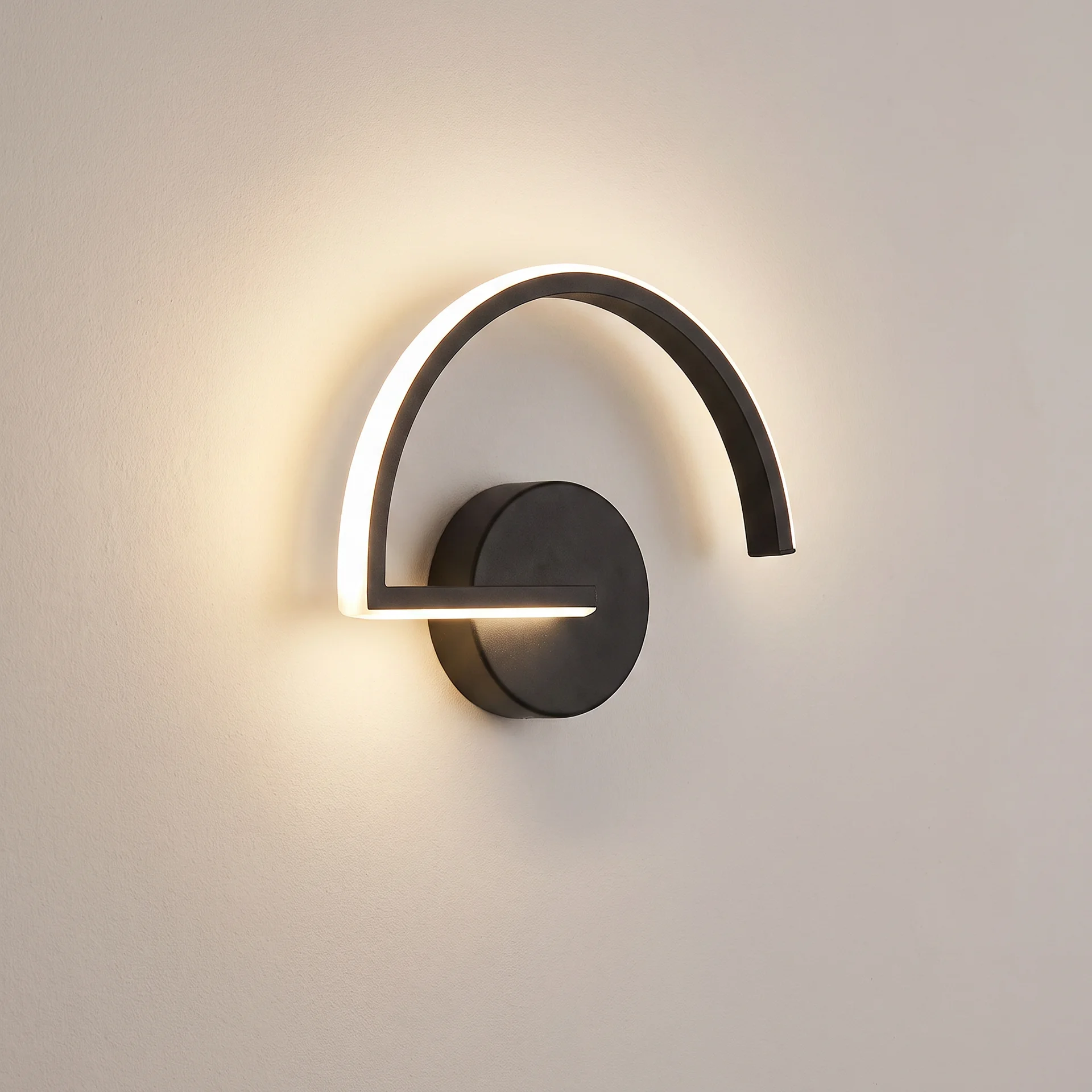 A modern wall-mounted light fixture with a matte black finish, featuring a curved backplate and a horizontal bar with a glowing white LED light strip, mounted on a warm off-white wall.
