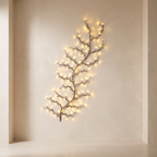A decorative branch-shaped wall light with warm white fairy lights, mounted on a beige plaster wall in a minimalist interior setting.