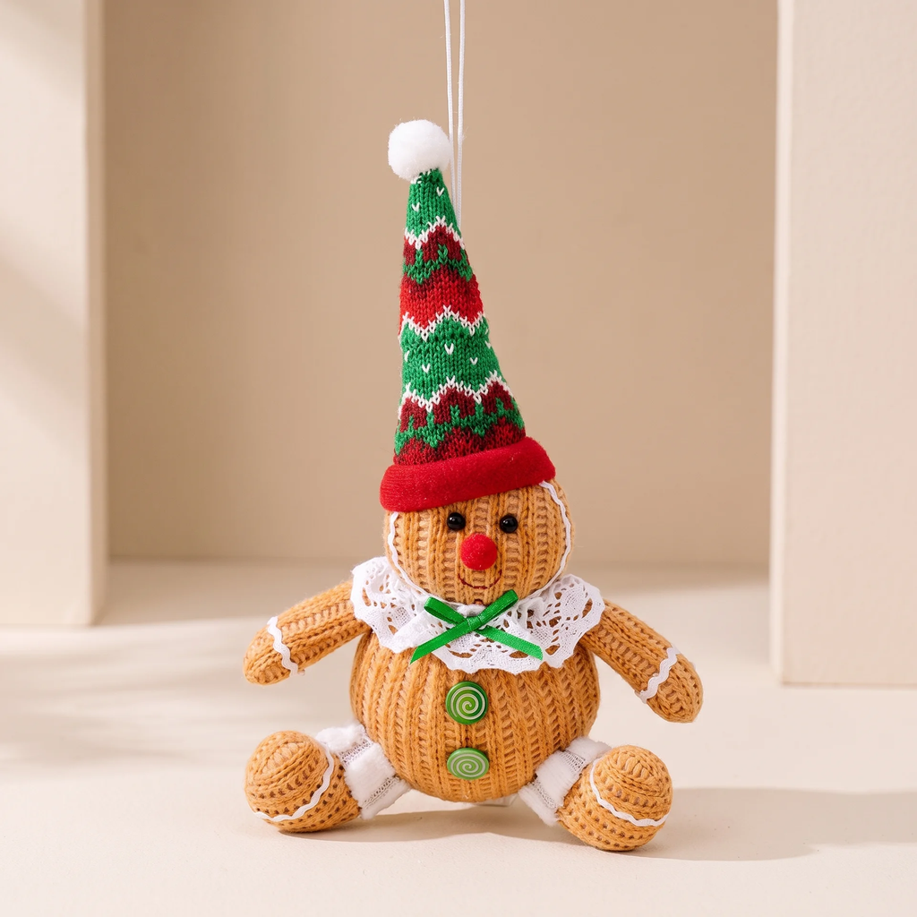 A knitted gingerbread man Christmas ornament hanging against a warm beige background. The ornament features a brown textured body, white lace collar, green buttons, and a red and green patterned knit hat with a white pom-pom.