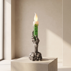 A decorative candle holder shaped like a skeletal hand holding a green candle with a glowing flame, displayed on a stone pedestal in a minimalist beige room.
