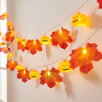 Halloween string lights featuring glowing orange jack-o'-lantern pumpkins and red-orange maple leaves with printed faces, shown lit against a warm off-white wall.
