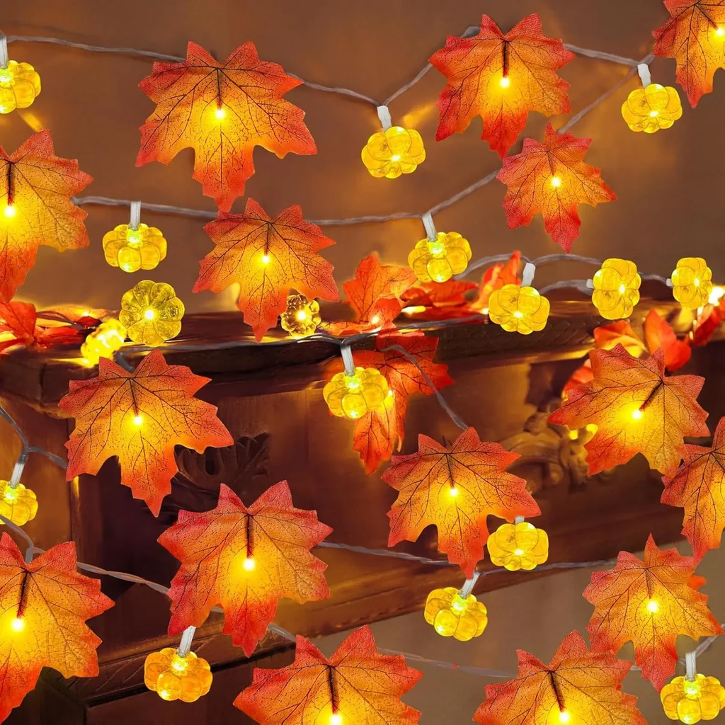 Warm white LED string lights featuring realistic orange and red autumn maple leaves and glowing pumpkin-shaped bulbs, draped over a dark wooden surface.