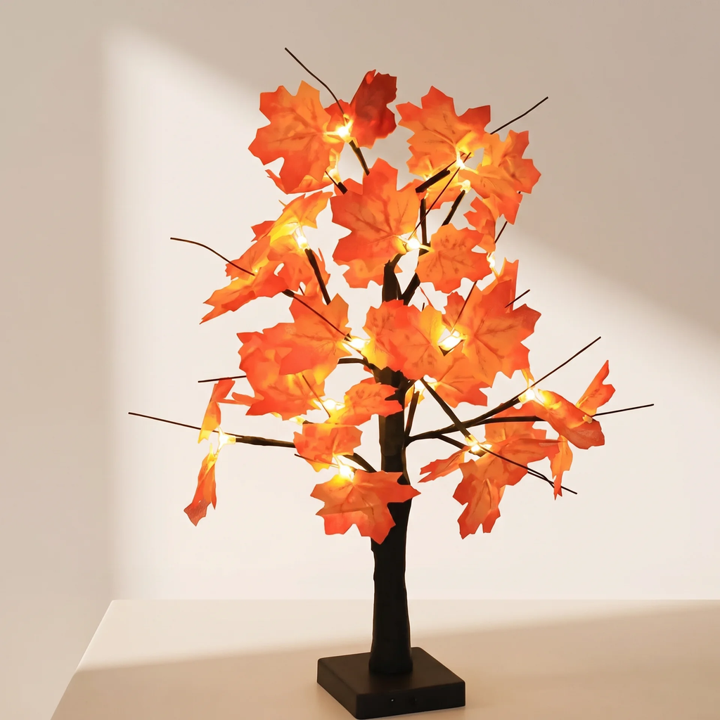 A decorative artificial tree with glowing orange maple leaves and warm white string lights, mounted on a black trunk and square base against a plain light wall.