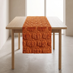 An orange table runner with a 3D pumpkin pattern, draped over a light wood dining table in a minimalist room with beige walls and concrete floors.