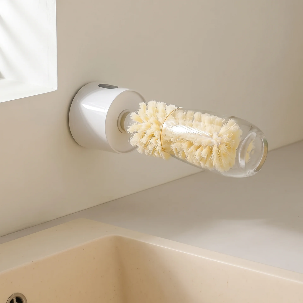 A wall-mounted glass bottle brush with cream-colored bristles, shown attached to a bathroom wall above a sink.