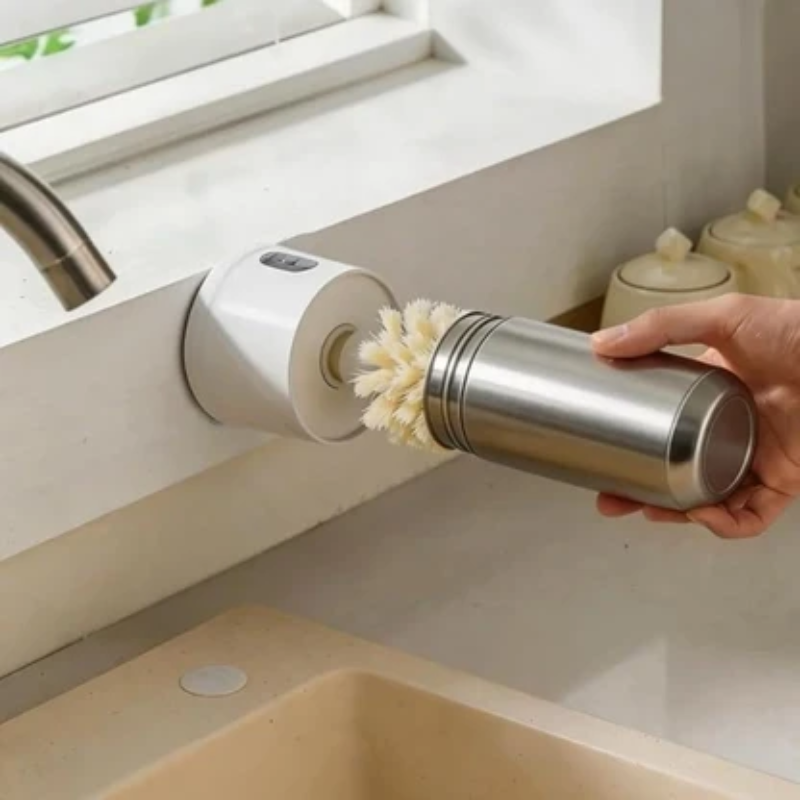 A stainless steel cylindrical cleaning brush with white bristles is being attached to a white wall-mounted faucet holder in a kitchen sink area. A hand is holding the brush, and the sink, faucet, and countertop are visible.