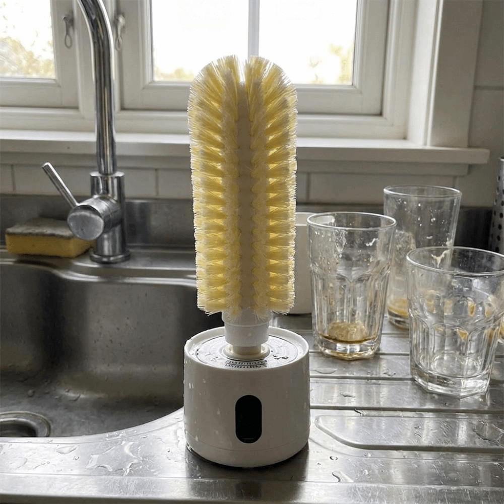 A tall yellow-handled bottle brush standing upright on a white cylindrical base on a stainless steel kitchen sink counter, with glasses and a sink visible in the background.
