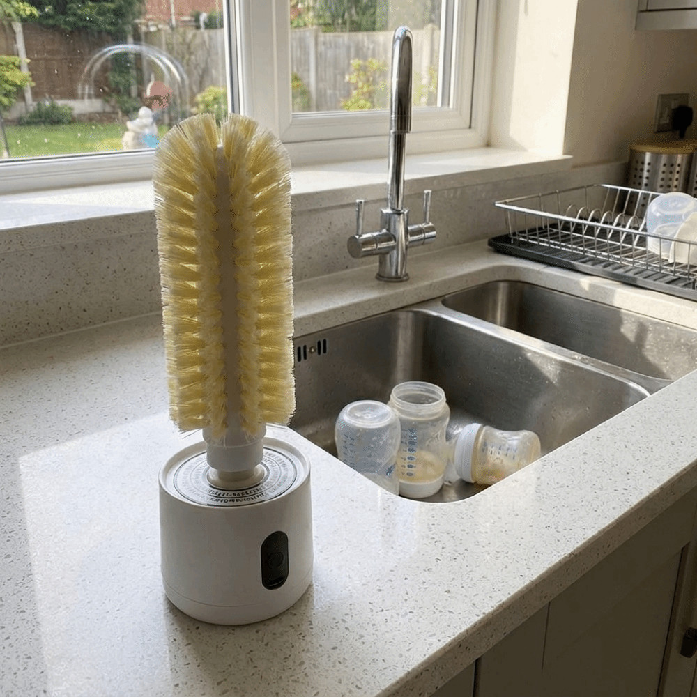 A white electric baby bottle cleaning brush with beige bristles stands on a speckled kitchen countertop next to a sink filled with baby bottles.