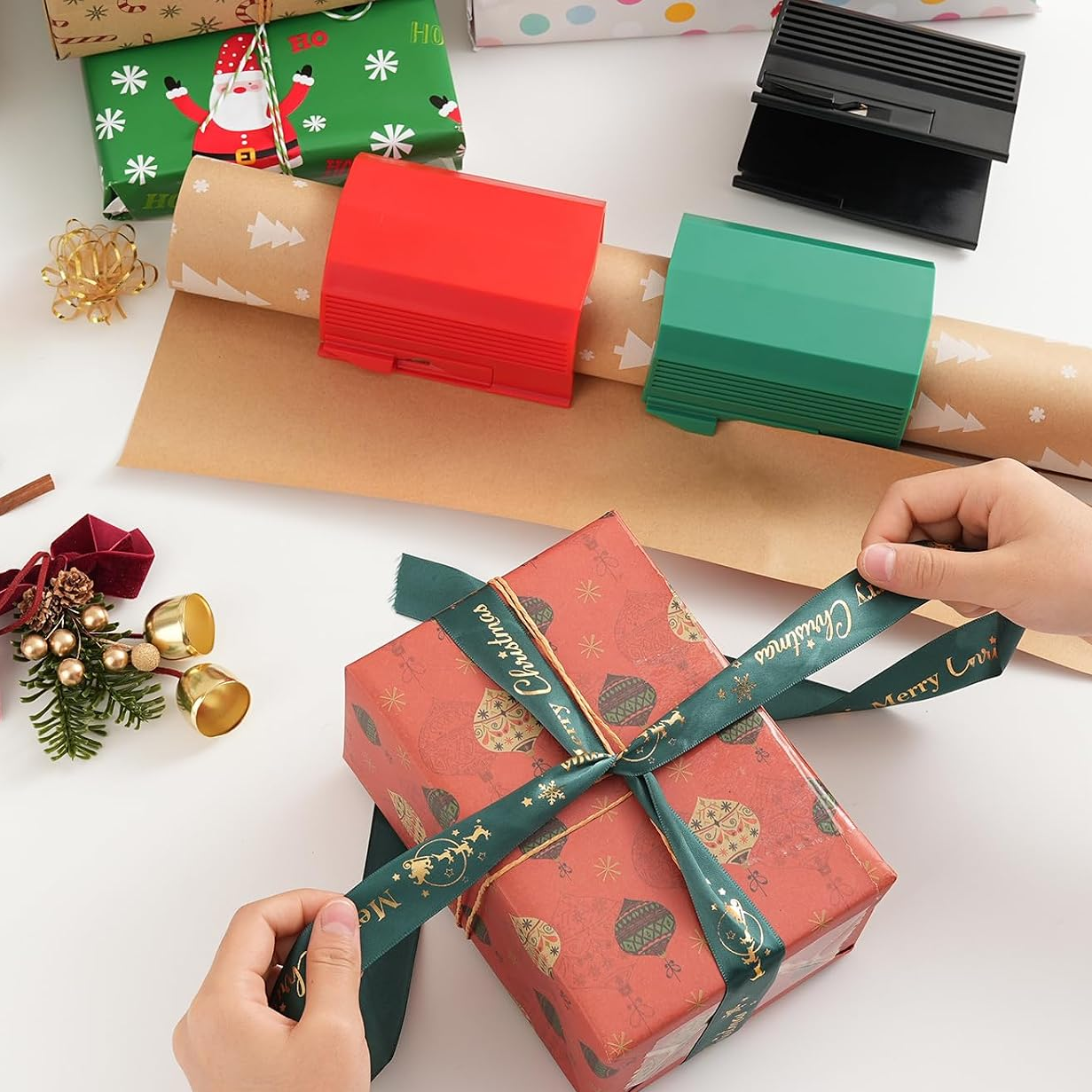 Person wrapping a gift with red paper and green ribbon on a white surface with Christmas decorations.