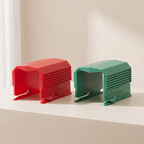Two small plastic accessory pieces, one red and one green, displayed side by side on a white surface against a warm off-white background.
