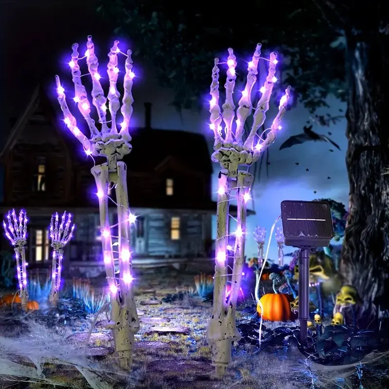Halloween yard decorations featuring two large illuminated skeleton hands with glowing purple lights, set in a spooky garden scene at night with a house, tree, pumpkins, and cobwebs.