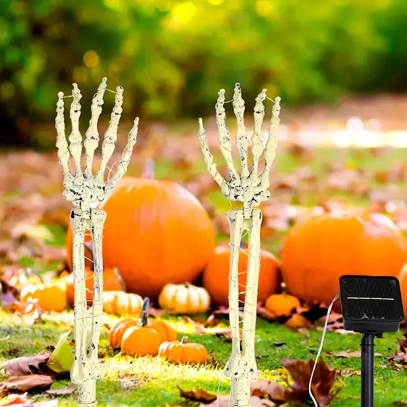 Two skeletal hand garden stakes stand on grass outdoors, with pumpkins and autumn leaves blurred in the background. A small solar panel on a stand is attached by a white cord.
