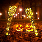 Two skeleton hand Halloween yard lights with warm white string lights stand on either side of two glowing jack-o'-lantern pumpkins on a leaf-covered ground at night.