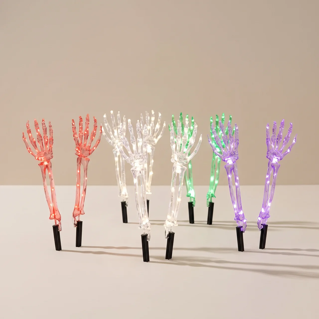 Six decorative skeletal hand garden lights in different colors (red, white, green, and purple) with black stakes, arranged on a light beige surface against a neutral beige background.