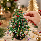 A hand is adjusting a small, glittery green Christmas tree decorated with colorful rhinestone ornaments, set against a warm, softly blurred holiday background.