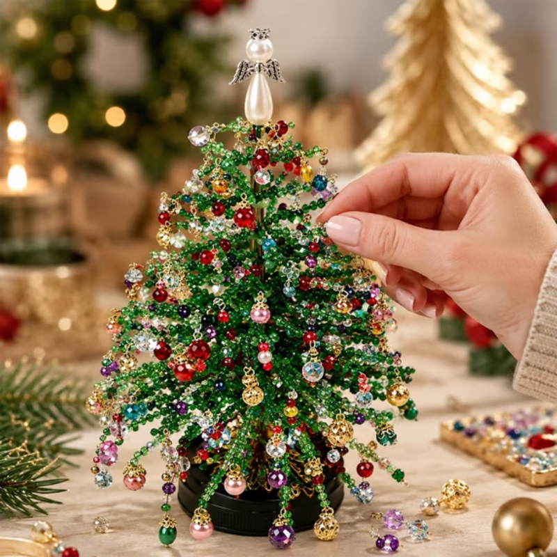 A hand is adjusting a small, glittery green Christmas tree decorated with colorful rhinestone ornaments, set against a warm, softly blurred holiday background.