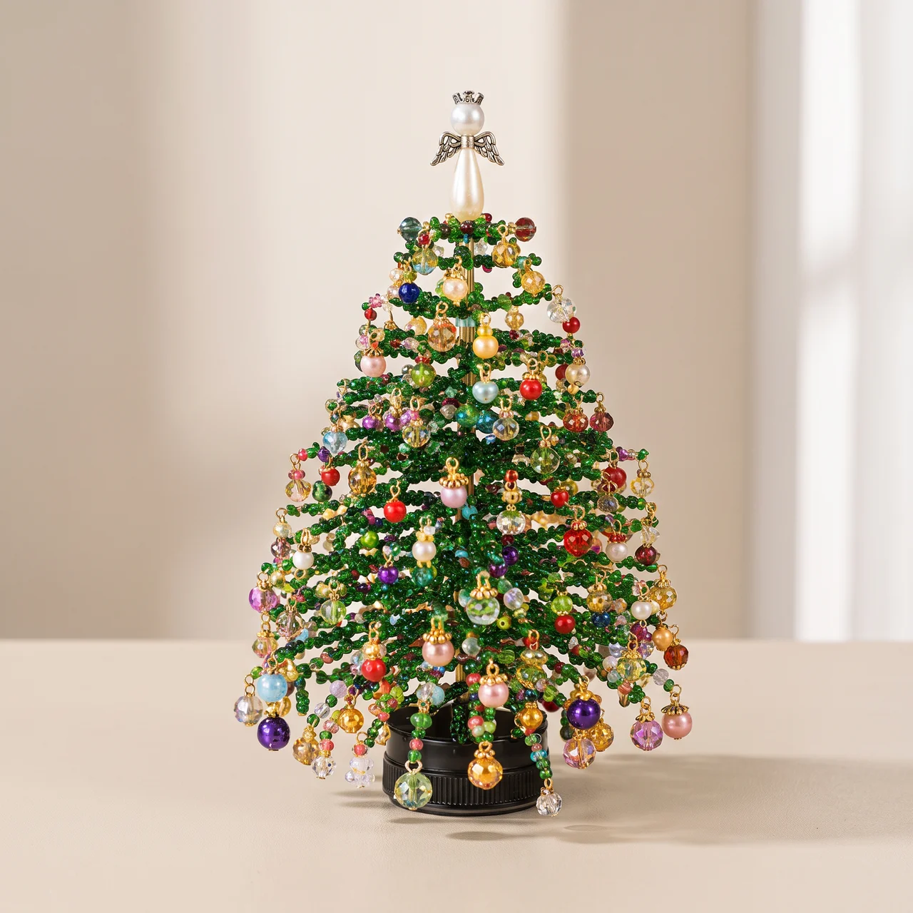 A miniature green Christmas tree decorated with colorful glass ornaments, hanging beads, and a silver angel topper, displayed on a light beige surface against a soft neutral background.