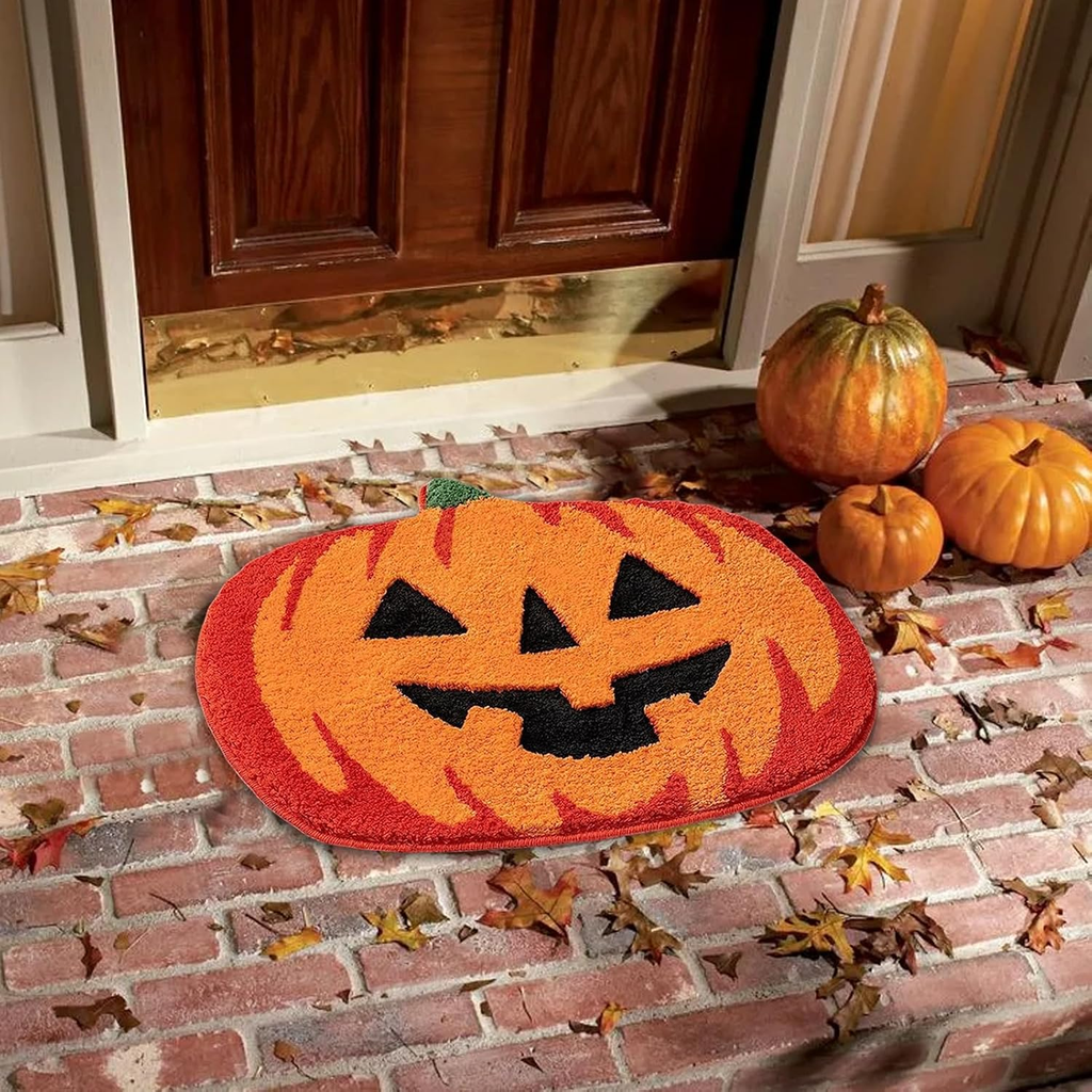 An oval Halloween doormat with a jack-o'-lantern face design in orange, red, and black, placed on a brick doorstep next to pumpkins and scattered autumn leaves.